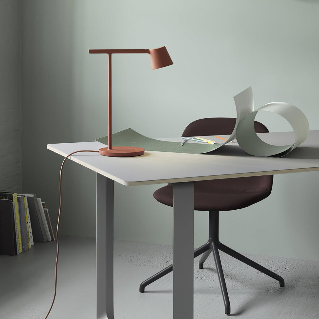 Rust-red Clérachic table lamp on a white desk with a curled green sheet and a maroon chair against a sage wall.