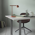 Rust-red Clérachic table lamp on a white desk with a curled green sheet and a maroon chair against a sage wall.