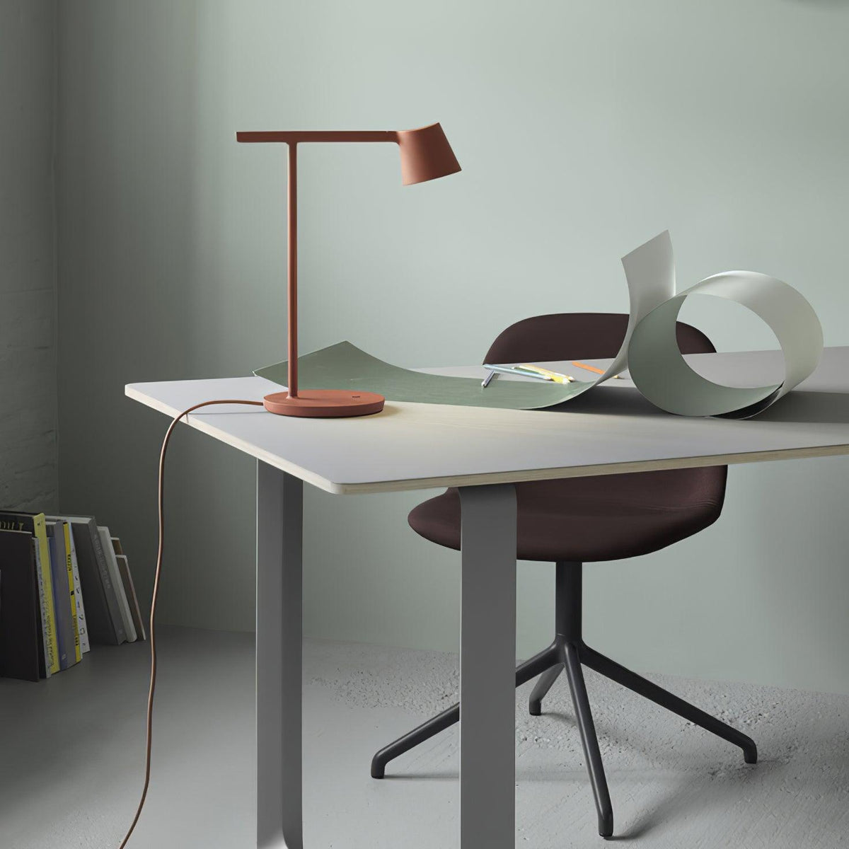 Rust-red Clérachic table lamp on a white desk with a curled green sheet and a maroon chair against a sage wall.