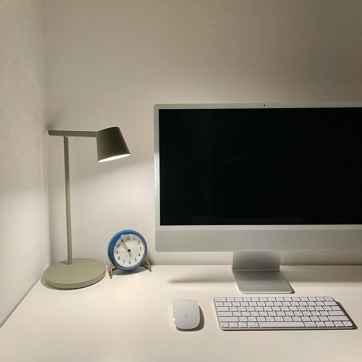 This Clérachic table lamp in olive, switched on beside a silver desktop computer and a small blue clock on a white desk.