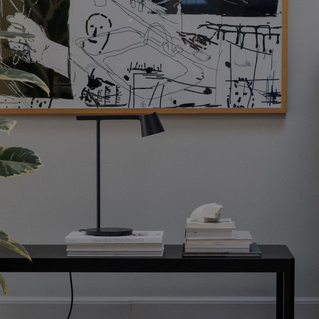 A black Clérachic table lamp standing on a stack of books on a black console, in front of a framed line drawing.