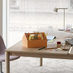A pale Clérachic table lamp in the background of a wooden table that holds an orange desk organiser and glasses.