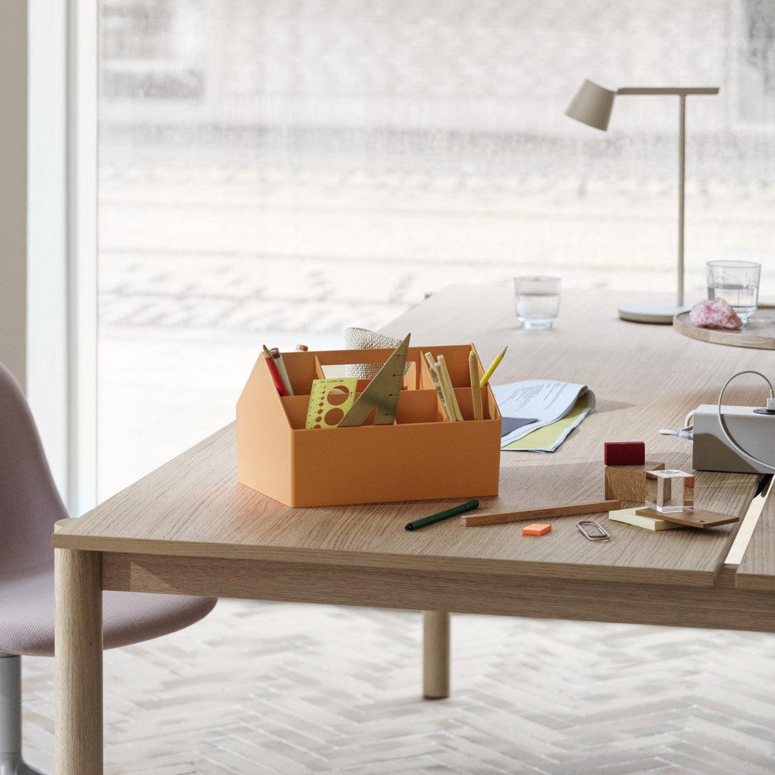 A pale Clérachic table lamp in the background of a wooden table that holds an orange desk organiser and glasses.