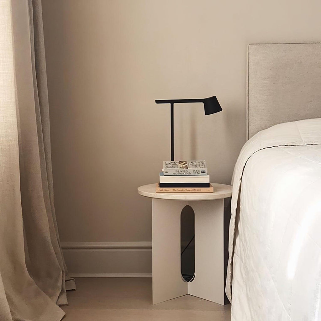 A black Clérachic table lamp on a small round bedside table with a pile of books, next to a made bed and beige curtain.