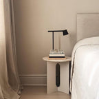 A black Clérachic table lamp on a small round bedside table with a pile of books, next to a made bed and beige curtain.