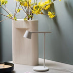 White Clérachic table lamp beside a tall ribbed cream vase of yellow blossom, with a grey-green wall behind.