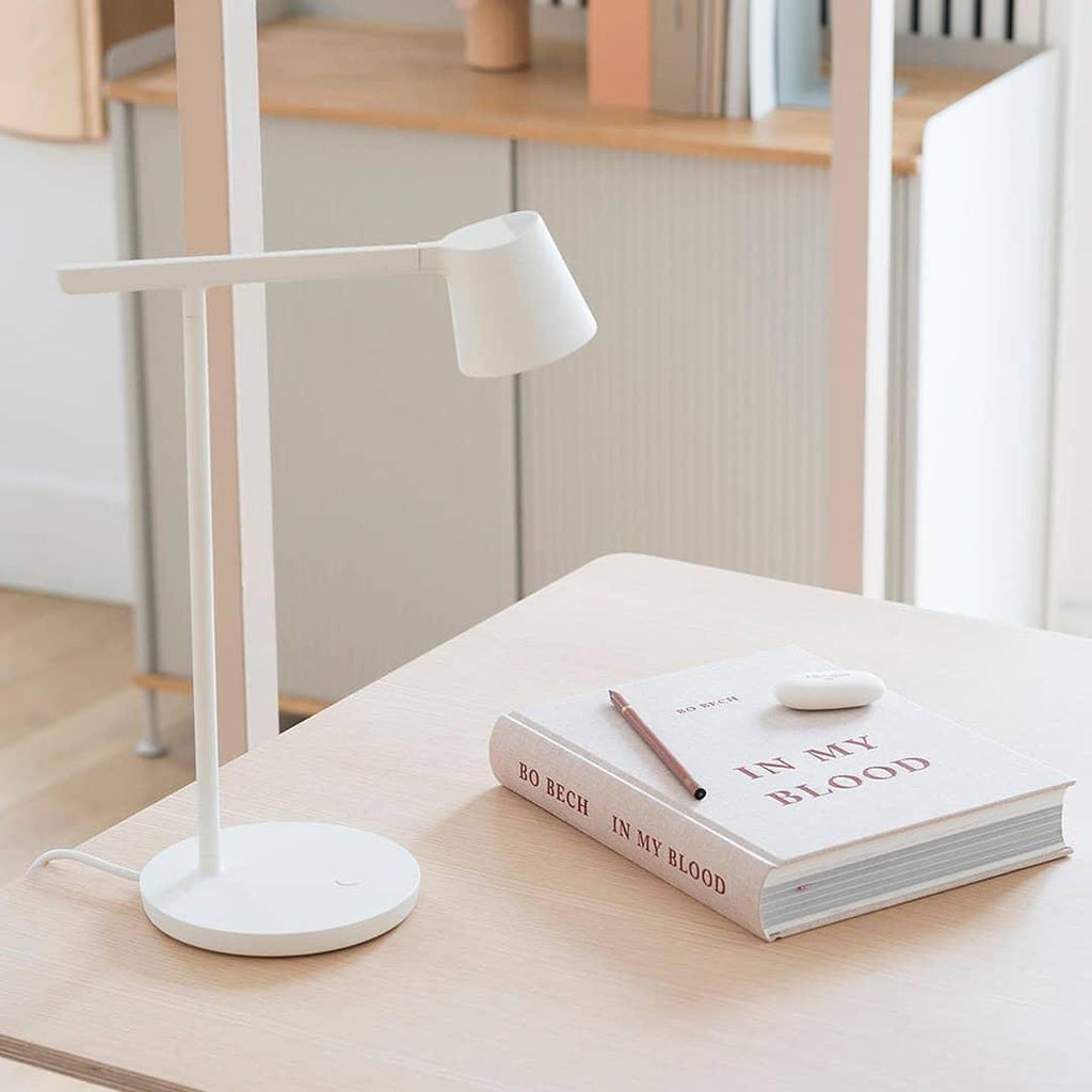 This Clérachic table lamp in white on a pale wood desk, next to a closed book with a pencil on top.