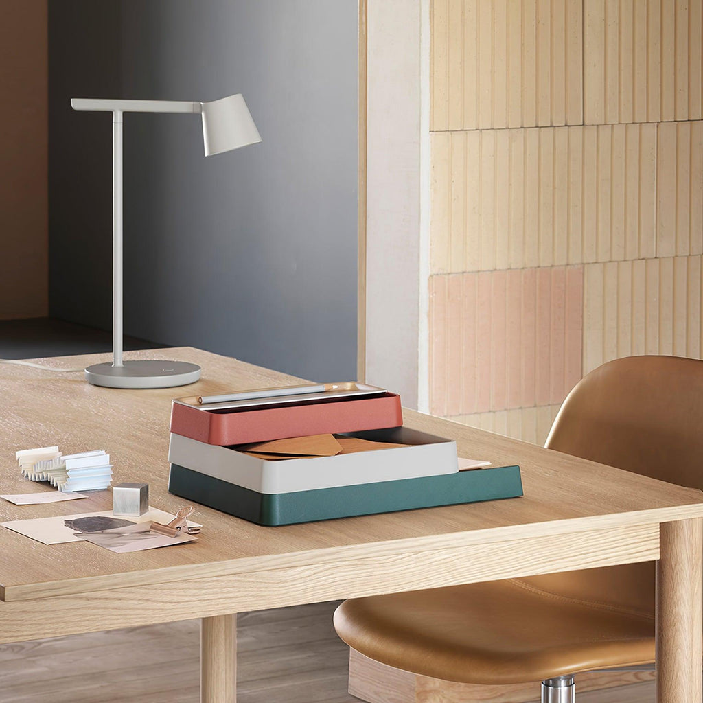 A pale-grey version of this Clérachic table lamp on a wooden desk with stacked boxes and a tan chair behind.
