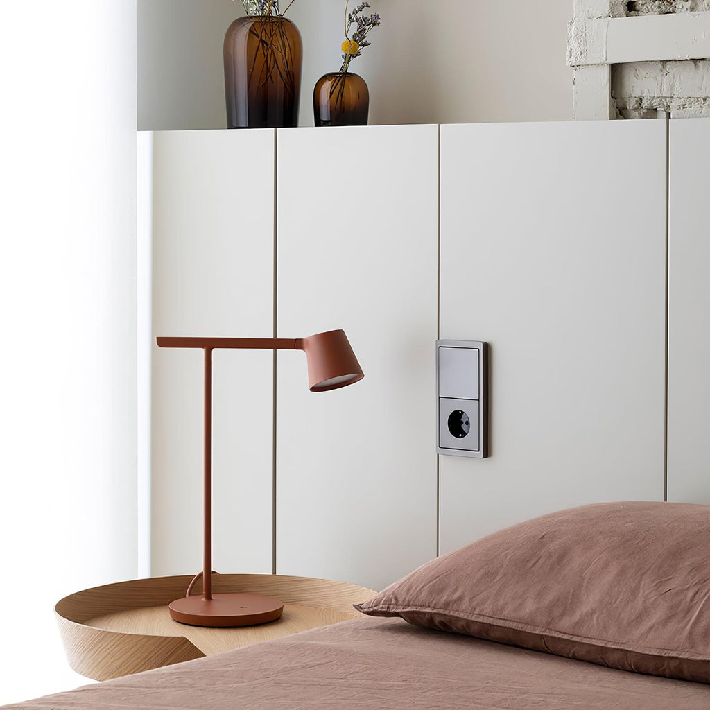 Rust-red Clérachic table lamp on a wooden tray resting on a bed, beside a white cabinet with a wall socket.