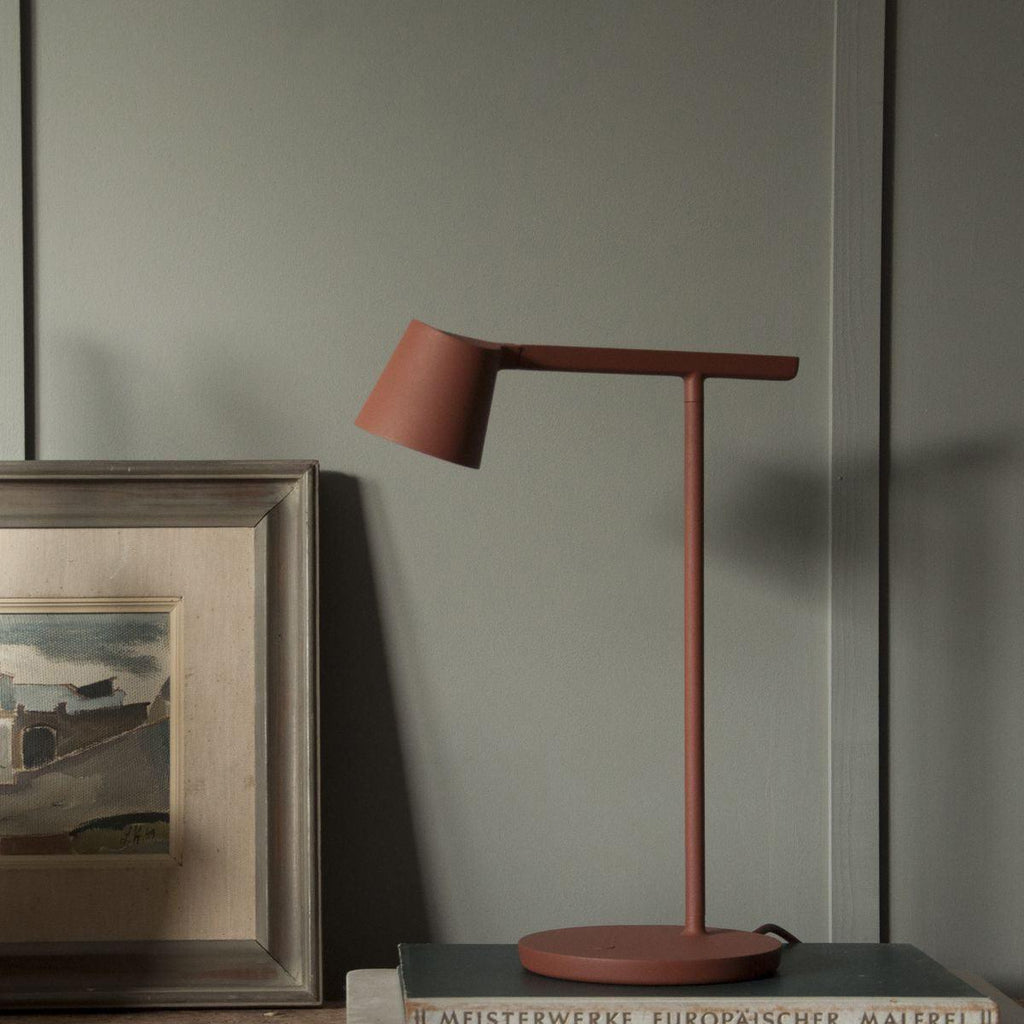 Rust-red Clérachic table lamp on a shelf in front of a green-grey wall, leaning near a framed landscape painting.
