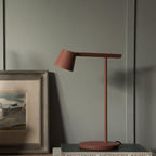 Rust-red Clérachic table lamp on a shelf in front of a green-grey wall, leaning near a framed landscape painting.