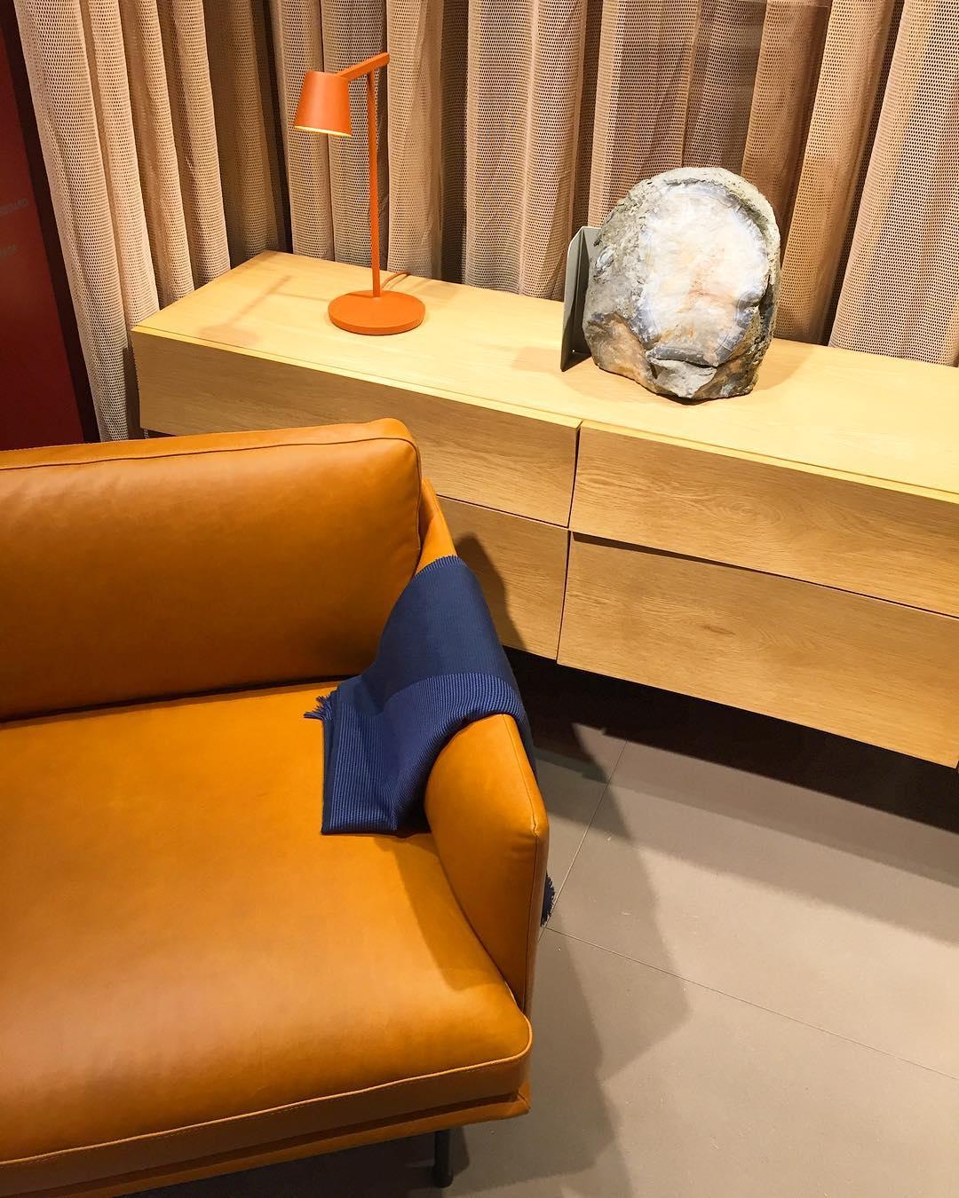 A small orange-red Clérachic table lamp on a pale wood sideboard, beside a tan leather sofa and gathered curtains.