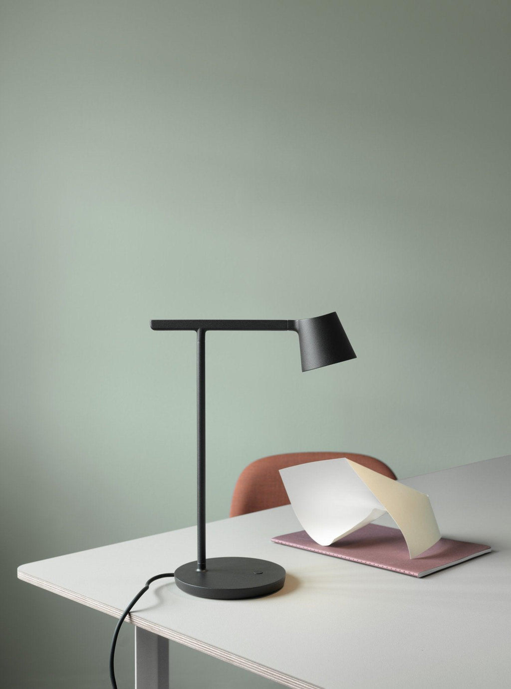 Black Clérachic table lamp on a white desk against a sage-green wall, with a pink notebook and paper at its side.