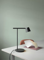 Black Clérachic table lamp on a white desk against a sage-green wall, with a pink notebook and paper at its side.
