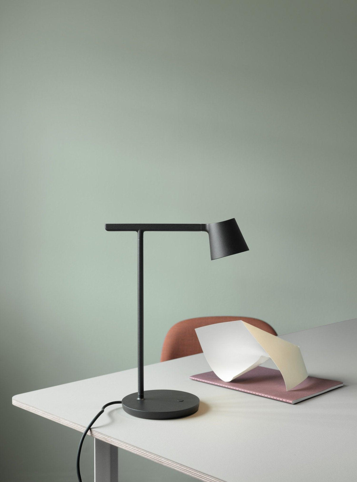 Black Clérachic table lamp on a white desk against a sage-green wall, with a pink notebook and paper at its side.