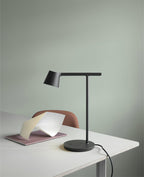 A black Clérachic table lamp seen from a low angle on a white desk, with its cord trailing off and a sage wall behind.