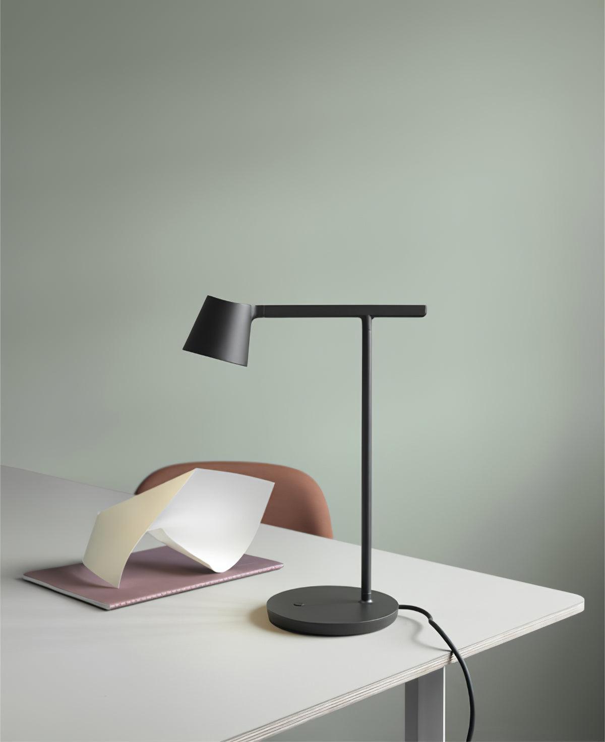 A black Clérachic table lamp seen from a low angle on a white desk, with its cord trailing off and a sage wall behind.