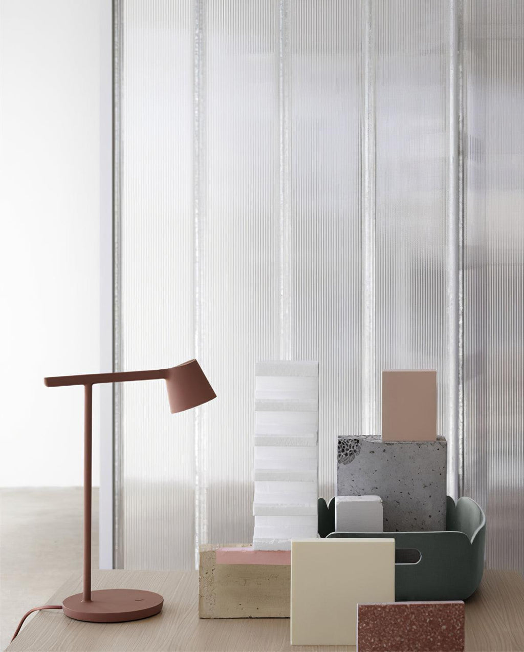 Rust-red Clérachic table lamp next to stacked pastel blocks and a green tray, in front of a ribbed glass wall.