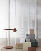 Rust-red Clérachic table lamp next to stacked pastel blocks and a green tray, in front of a ribbed glass wall.