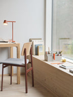 A rust-red Clérachic table lamp on a small desk by a window, with a wooden chair and a shelf of books to one side.