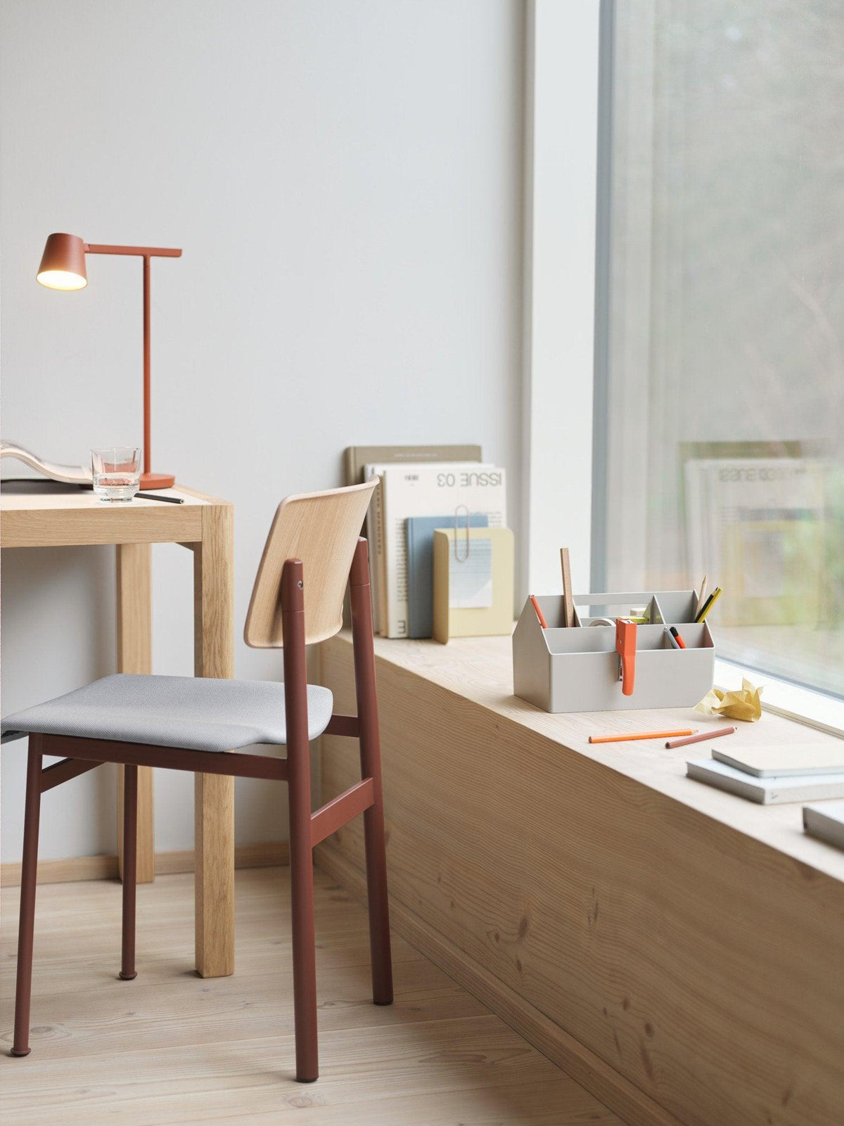 A rust-red Clérachic table lamp on a small desk by a window, with a wooden chair and a shelf of books to one side.