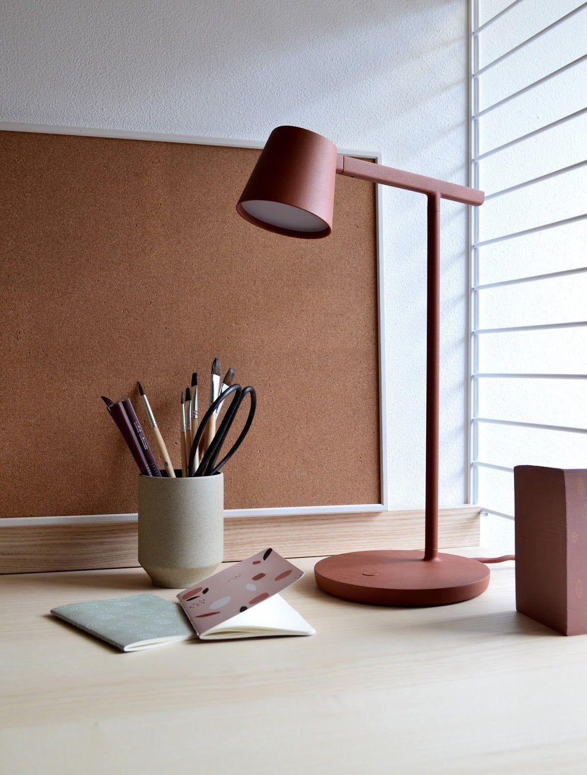 Rust-red Clérachic table lamp switched on beside a pot of pencils and a cork board, with window blinds at the right.