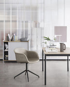A white Clérachic table lamp on a distant desk in a studio, with a grey upholstered chair in the foreground.