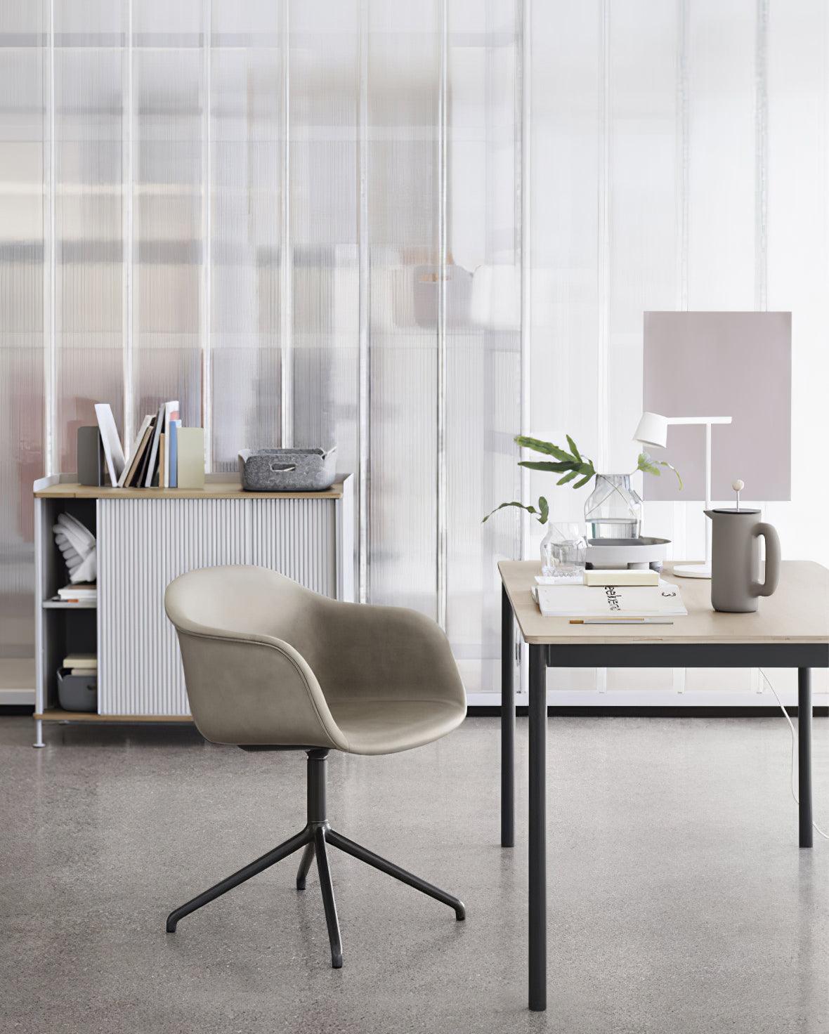 A white Clérachic table lamp on a distant desk in a studio, with a grey upholstered chair in the foreground.