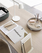 Looking down on a white Clérachic table lamp on a pale wood desk with an open magazine, a tray and a pot of pencils.