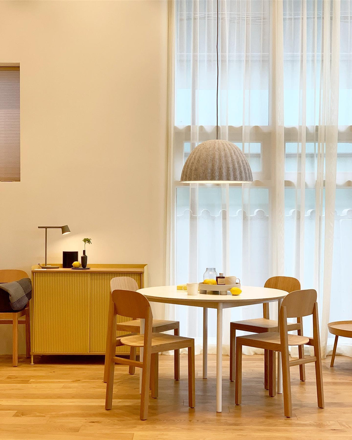A small Clérachic table lamp on a yellow sideboard in a warm room with a round dining table and a fabric pendant above.