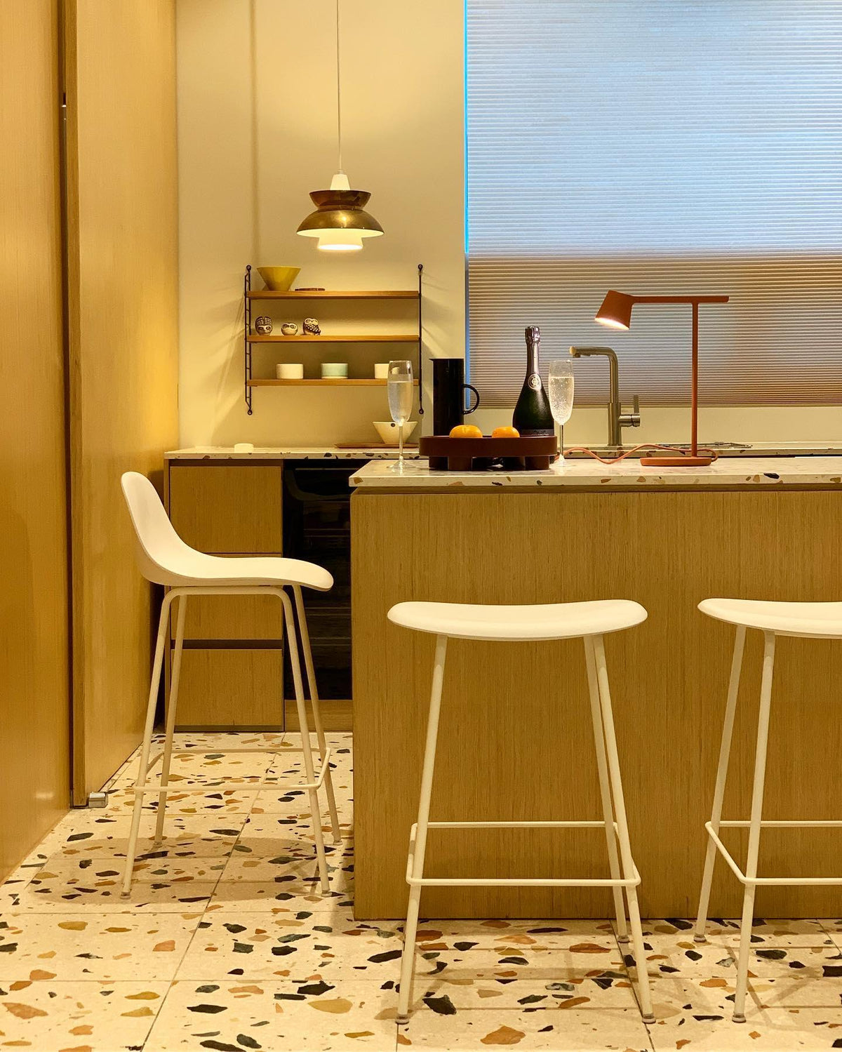 Rust-red Clérachic table lamp on a kitchen counter, with white bar stools and a terrazzo floor in front.