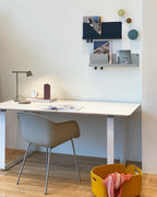 An olive Clérachic table lamp on a white desk with a clock, a grey chair and wall shelves above.