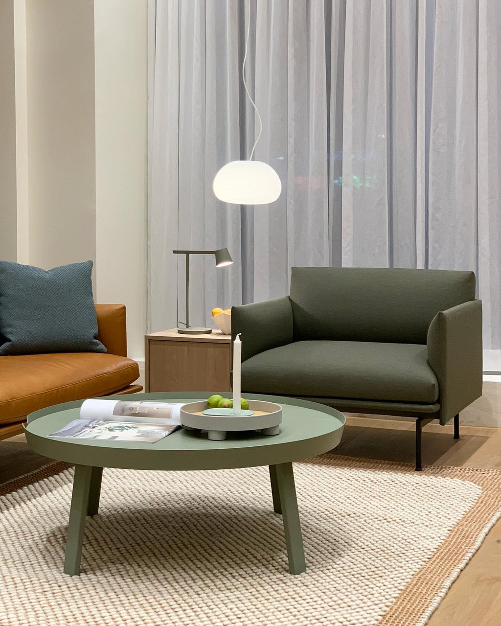 A Clérachic table lamp on a side table in a living room with a green armchair, a round green coffee table and a white pendant.