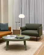 A Clérachic table lamp on a side table in a living room with a green armchair, a round green coffee table and a white pendant.