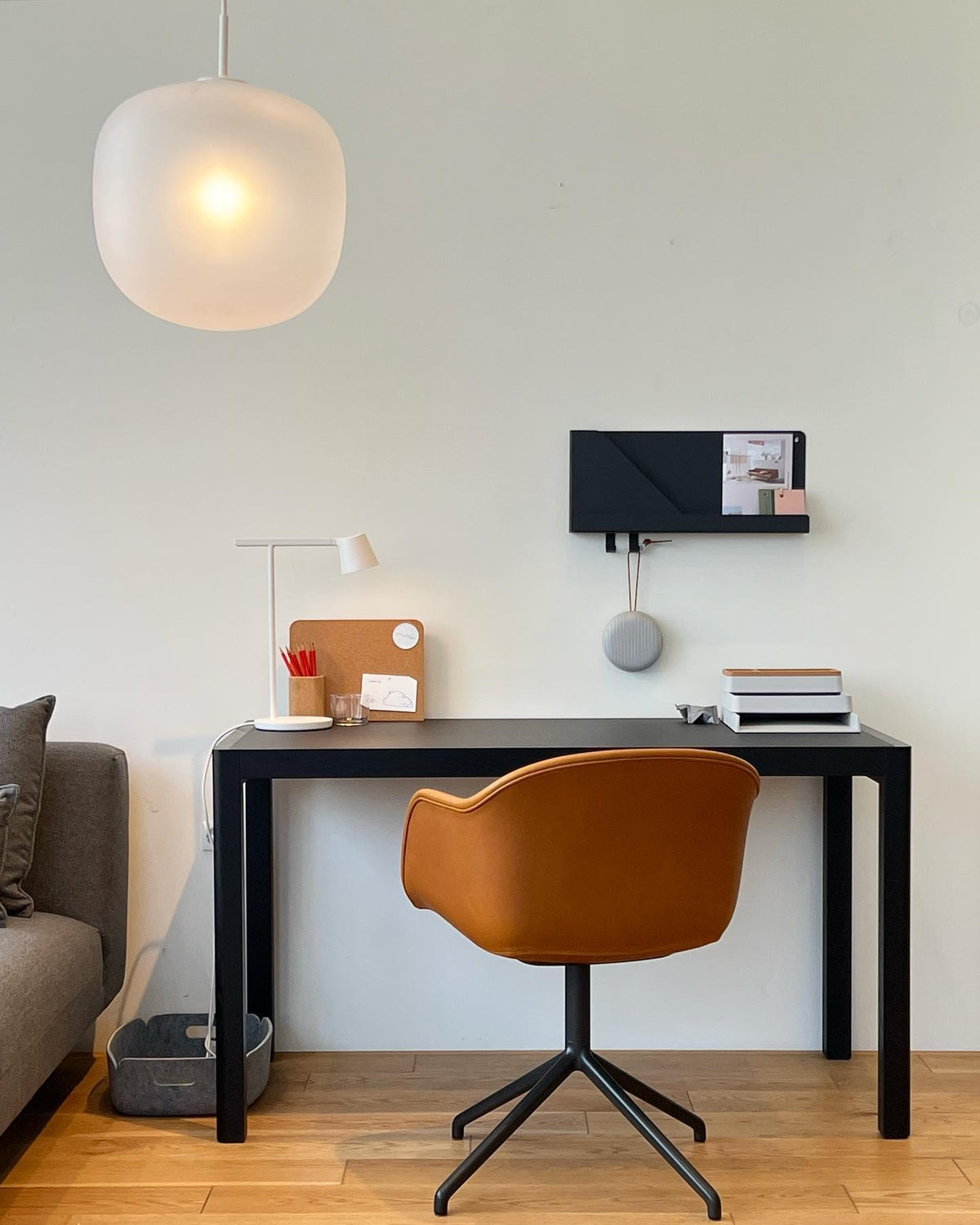 A white Clérachic table lamp on a black desk with a tan chair, a wall shelf and a frosted pendant overhead.