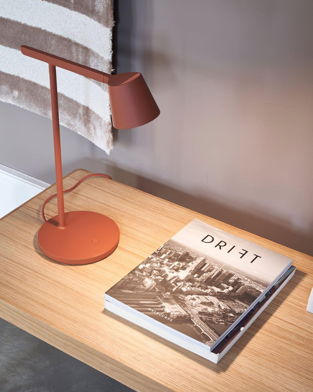 Rust-red Clérachic table lamp on a pale wood desk, with a large photo book lying open in front of the round base.