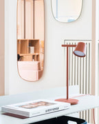 A rust-red Clérachic table lamp switched on over a white desk with magazines, with a tall mirror on the wall behind.