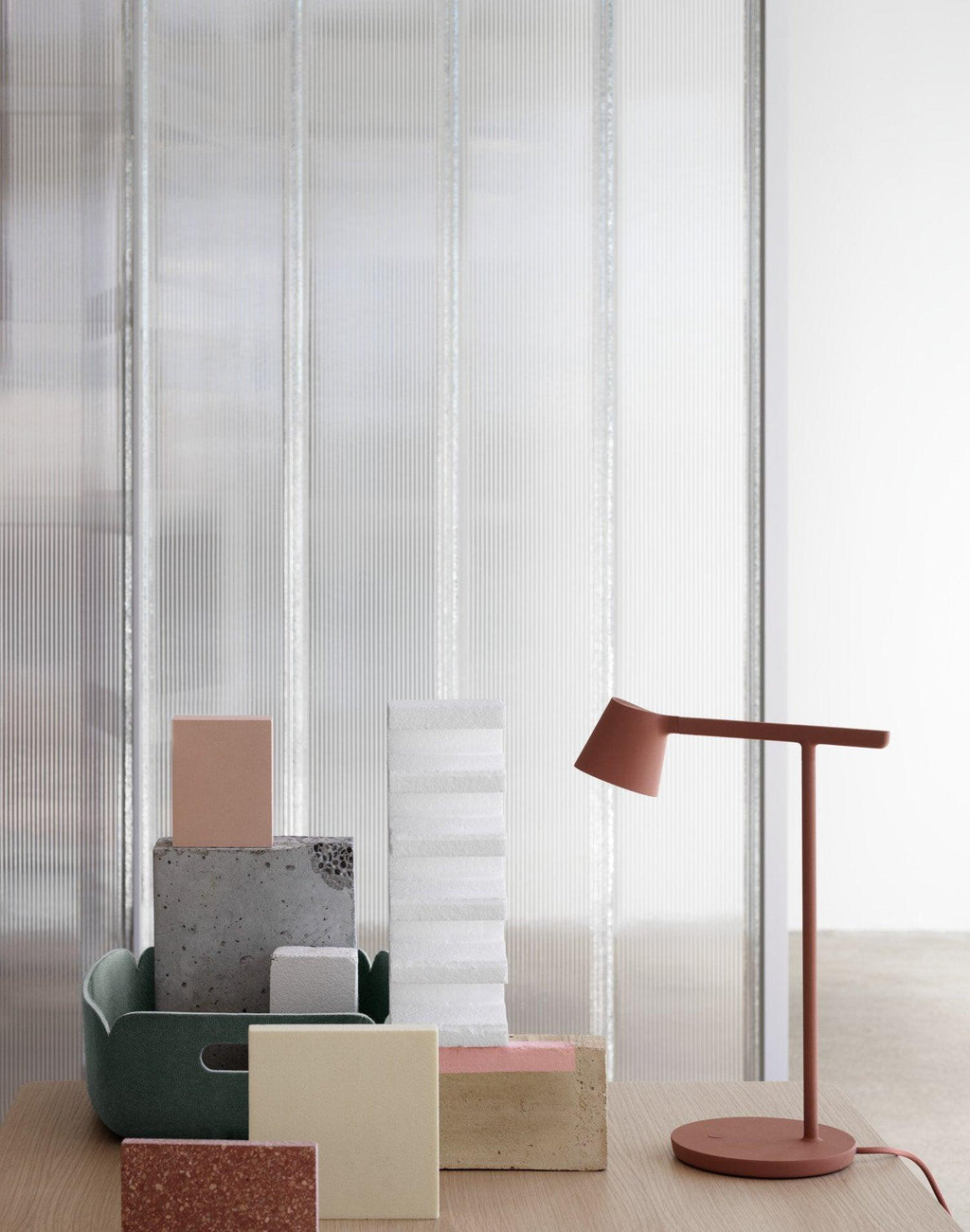 Rust-red Clérachic table lamp beside pastel blocks and a dark green tray, in front of a ribbed glass wall.