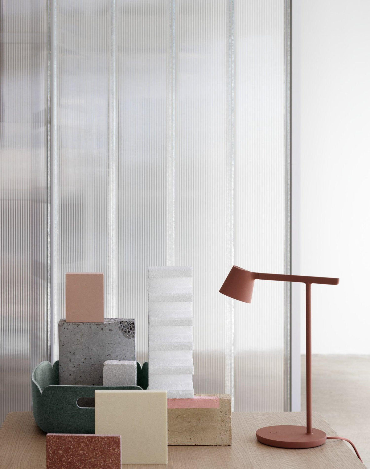 Rust-red Clérachic table lamp beside pastel blocks and a dark green tray, in front of a ribbed glass wall.