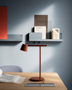 A rust-red Clérachic table lamp on a wooden desk against a blue-grey wall, below a shelf of small objects.