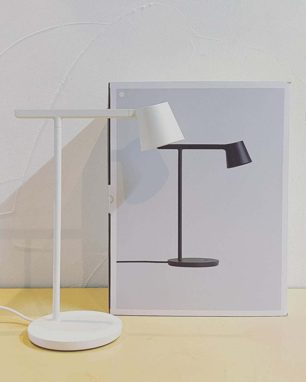 White Clérachic table lamp on a pale yellow surface in front of a printed poster that shows a black version.