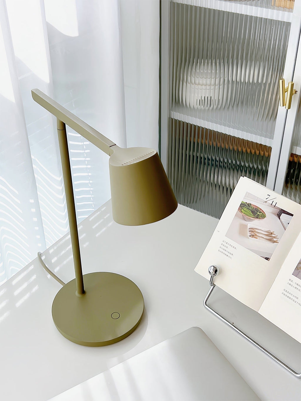 Olive Clérachic table lamp on a white desk beside an open recipe book, with sheer curtains and a glass cabinet behind.