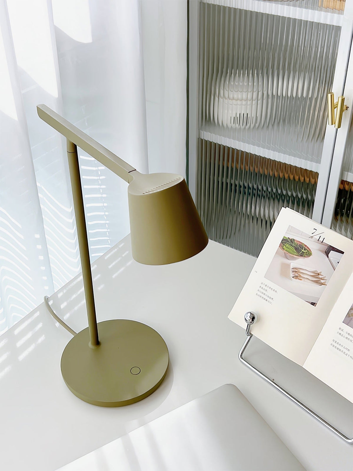 Olive Clérachic table lamp on a white desk beside an open recipe book, with sheer curtains and a glass cabinet behind.