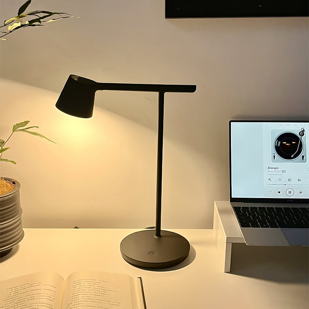 This Clérachic table lamp in black lit warmly on a desk, with a laptop at its side and an open notebook in front.