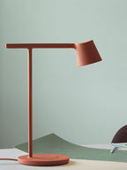 A rust-red Clérachic table lamp close up against a pale green wall, with a green curved sheet behind the base.