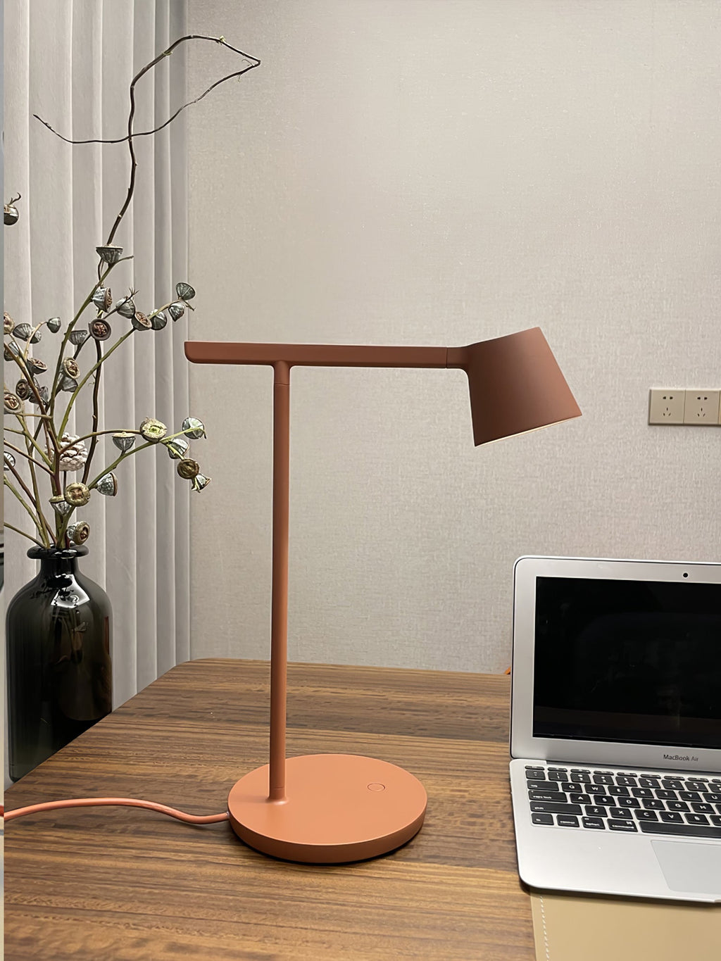 A rust-red Clérachic table lamp on a wooden desk next to a laptop and a vase of dried stems.