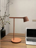 A rust-red Clérachic table lamp on a wooden desk next to a laptop and a vase of dried stems.