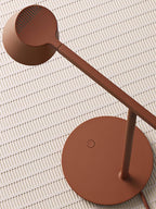 Overhead close view of a rust-red Clérachic table lamp, with its round head and base on a ribbed pale tile.