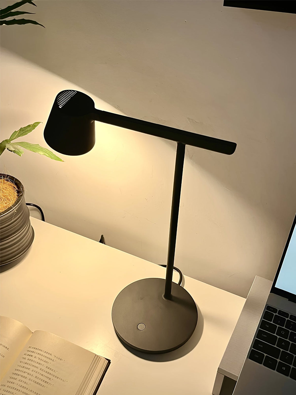 Black Clérachic table lamp glowing on a white desk, with a laptop and an open book beside the round base.