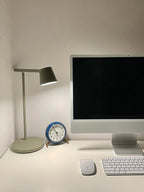 A grey-green Clérachic table lamp beside a desktop computer, a keyboard and a blue clock on a white desk.